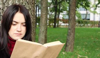 ritratto di una bella giovane donna che è seduta sotto un albero e sta leggendo il suo libro preferito in un parco cittadino sull'erba verde in una piacevole giornata di sole. concetto di vacanza, istruzione e studio. foto