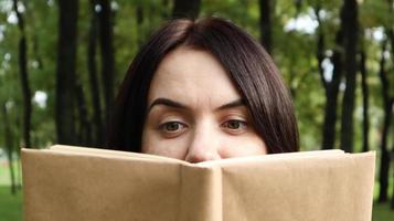 ritratto di una giovane donna con un libro aperto senza titolo tenuto vicino al viso. bruna che copre metà del viso con un libro nel parco in una giornata di sole. concetto di educazione e persone, messa a fuoco selettiva. foto