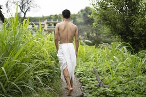 giovane ragazzo indiano in forma, camminando su un sentiero accanto ai raccolti nel campo. un sacerdote indiano che cammina indossando dhoti bianchi. uomo religioso indiano. foto