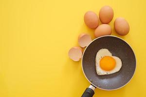 uovo fritto a forma di cuore in padella con guscio d'uovo e uova di gallina su sfondo giallo. vista dall'alto. concetto di cibo sano. copia spazio per il testo foto