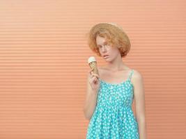 giovane donna rossa riccia allegra con cappello di paglia e prendisole blu che mangia il gelato su sfondo beige. divertimento, estate, moda, concetto di gioventù foto