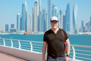 Ritratto di uomo turistico sorridente europian caucasico rosso con occhiali e cappello impressionato nella moderna dubai, emirati arabi uniti sul paesaggio urbano, sullo skyline e sullo sfondo del mare in una giornata di sole foto