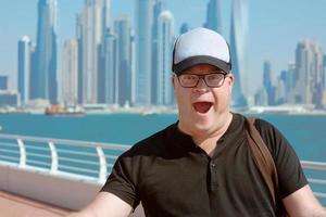 Ritratto di uomo turistico sorridente europian caucasico rosso con occhiali e cappello impressionato nella moderna dubai, emirati arabi uniti sul paesaggio urbano, sullo skyline e sullo sfondo del mare in una giornata di sole foto