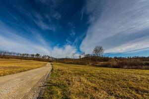 strada sterrata e una collina con boschi e nuvole fini al cielo foto