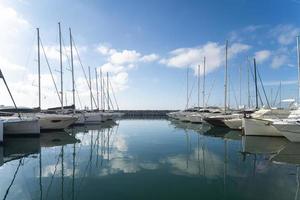 marina con barche che si riflettono sul mare e cielo azzurro con nuvole bianche foto