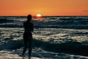 silhouette femminile nelle onde del mare blu al tramonto estivo, mezzo sole sotto l'orizzonte, vacanza sulla spiaggia foto