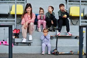 giovane madre alla moda con quattro bambini seduti sul podio sportivo allo stadio, mangia mela e beve acqua. la famiglia trascorre il tempo libero all'aria aperta con scooter e pattini. foto