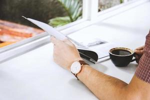 primo piano della mano dell'uomo che tiene una rivista e una tazza di caffè sul tavolo bianco foto