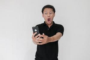 wow il viso del tuo uomo asiatico ha scioccato ciò che vede nello smartphone su sfondo grigio isolato. indonesia uomo indossare camicia nera isolato sfondo grigio foto