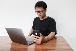 il giovane asiatico è confuso e scioccato quando chiama al telefono con il laptop accanto. uomo indonesiano che indossa una camicia blu foto