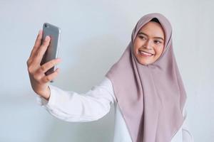 la giovane donna asiatica dell'islam che indossa il velo sorride e si fa selfie con il suo telefono cellulare. donna indonesiana su sfondo grigio foto