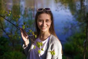 una ragazza con occhiali da sole vicino al lago, con in mano un ramo di un albero foto
