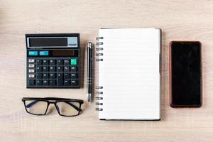 vista piana e dall'alto degli accessori per uomo d'affari sul desktop in legno dell'ufficio con calcolatrice, occhiali, penna, libro e smartphone foto