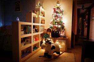 bambina sulla mucca a dondolo giocattolo contro l'albero di natale con ghirlande brillanti sulla sera a casa. foto