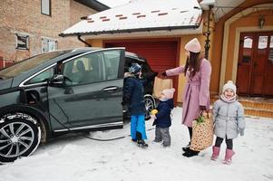 giovane donna con bambini tiene borse ecologiche e carica un'auto elettrica nel cortile di casa sua. foto