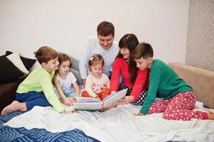 la grande famiglia felice si diverte insieme in camera da letto. concetto di mattina in famiglia numerosa. quattro bambini con i genitori indossano il pigiama e leggono un libro a letto a casa. foto