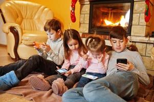 quattro bambini felici che guardano nel loro telefono cellulare a casa vicino a un caminetto nel caldo soggiorno in una giornata invernale. foto