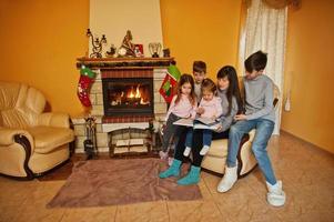 felice giovane famiglia numerosa accanto a un camino nel caldo soggiorno in una giornata invernale. madre con quattro bambini a casa legge il libro. foto