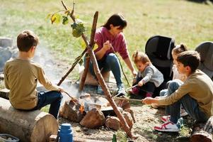 falò di famiglia in montagna. friggere le salsicce. madre con quattro bambini in campeggio. escursione autunnale e clima del campo. scaldare e cuocere insieme vicino alla fiamma. foto