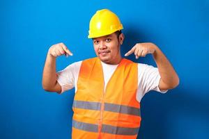 giovane uomo asiatico grasso su sfondo blu che indossa l'uniforme dell'appaltatore e il casco di sicurezza che punta e mostra con il dito foto