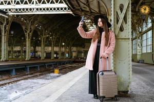 felice giovane donna sul binario della stazione ferroviaria in cappotto rosa e cappello nero foto