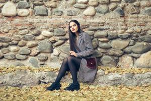 bella ragazza che indossa un cappotto invernale seduto sul pavimento di un parco urbano pieno di foglie autunnali. foto