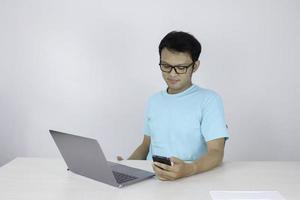 il giovane asiatico è serio e si concentra quando lavora su un telefono e un laptop sul tavolo. uomo indonesiano che indossa una camicia blu. foto