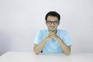 il giovane uomo asiatico indossa una camicia blu e occhiali con una faccia sorridente felice e tiene il mento sul tavolo. foto