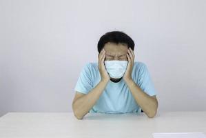 il giovane asiatico che indossa una maschera medica soffre di un forte mal di testa, premendo le dita sulle tempie, chiudendo gli occhi per alleviare il dolore con un'espressione facciale indifesa. foto