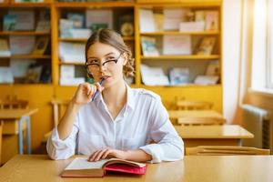 ragazza con gli occhiali seduta a un tavolo con un libro in classe, con una matita in mano, sguardo pensieroso foto