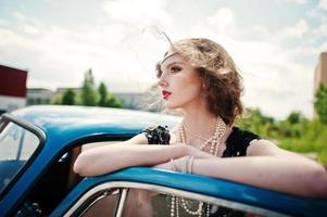 ritratto di bella modella riccia ragazza di moda con trucco luminoso in stile retrò appoggiato su una portiera di un'auto d'epoca. foto