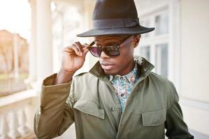 ritratto di moda di uomo afroamericano nero su mantello verde e cappello nero, resta sul balcone del palazzo giallo foto