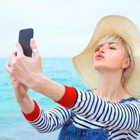 bella giovane donna caucasica bionda in vacanza con cappello di paglia, camicetta a righe e salopette di jeans che fanno selfie sullo smartphone sullo sfondo del mare blu incredibile foto