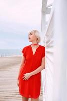 ritratto di bella donna bionda capelli corti piuttosto cool in abito di corallo rosso vicino a pergolato in legno bianco per la spiaggia foto