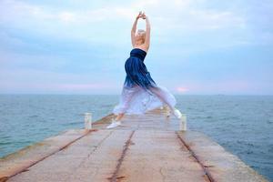 donna incinta bionda bianca caucasica con i capelli corti in abito lungo e scarpe da ginnastica felice di essere incinta e saltare in riva al mare all'alba foto