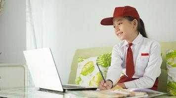 ragazza asiatica della scuola elementare che studia online a casa prendendo appunti dal computer portatile foto