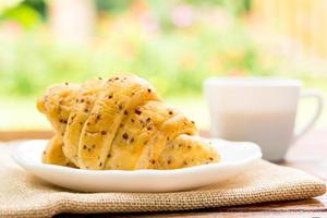 concetto di colazione. croissant con semi di perilla su piatto bianco e tazza bianca di caffè nero su tavola di legno con sfondo verde bokeh. foto