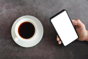 concetto di affari. caffè nero o espresso caldo in una tazza di ceramica bianca e tenere in mano lo schermo bianco su uno smartphone o un cellulare su un tavolo scuro. foto