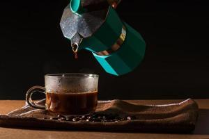 versare il caffè nero caldo dalla moka verde per pulire la tazza di caffè con fumo e chicchi di caffè su una tovaglia marrone e un tavolo di legno. beneficio del concetto di caffè. foto