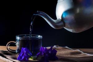 versare il tè di piselli farfalla da una pentola di metallo alla tazza con fiore viola su tovaglia marrone su sfondo nero. bevanda salutare da bere. erbe e concetto medico. foto