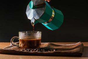 versare il caffè nero caldo dalla moka verde per pulire la tazza di caffè con fumo e chicchi di caffè su una tovaglia marrone e un tavolo di legno. beneficio del concetto di caffè.