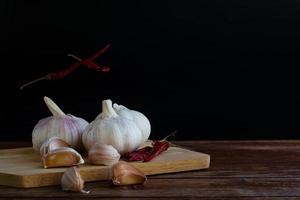 gruppo di aglio sul tagliere e alcuni spicchi d'aglio che galleggiano nell'aria e peperoncino rosso secco sul tavolo di legno con sfondo nero. copia spazio per il tuo testo. foto