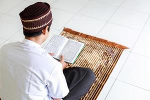 vista posteriore dell'uomo musulmano che tiene e legge il libro sacro al-quran sul tappeto di preghiera foto