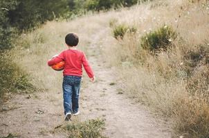 vista posteriore di un bambino con una palla in braccio che cammina lungo una strada foto