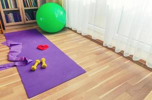 attrezzatura sportiva sul pavimento del soggiorno, tappetino da yoga viola, manubri gialli, fascia di resistenza in gomma rossa e palla aerobica fitness in gomma verde foto