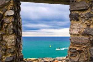 Vista dell'acqua del mare ligure, della scogliera rocciosa dell'isola di Palmaria e della barca attraverso la finestra del muro di pietra di mattoni, città costiera di portovenere foto