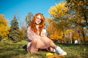 ritratti di un'affascinante ragazza dai capelli rossi con occhiali e un bel viso. ragazza in posa nel parco autunnale con un maglione e una gonna color corallo. foto