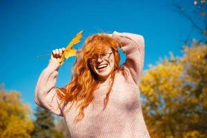 ritratti di un'affascinante ragazza dai capelli rossi con un viso carino. ragazza in posa nel parco autunnale con un maglione e una gonna color corallo. nelle mani di una ragazza una foglia gialla foto