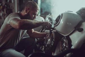 bell'uomo barbuto che ripara la sua moto in garage. un uomo che indossa jeans e una maglietta foto