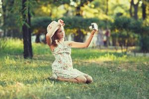 la bambina è giocata dalla macchina fotografica che si siede sull'erba nel parco. facendo selfie e fotografando il mondo intorno foto